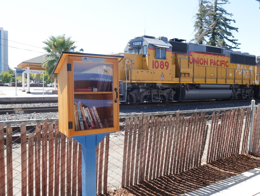A Little Free Library at RABA with two shelves of books sits in front of a fence, and a train track with a yellow Union Pacific train.