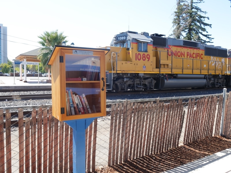 A Little Free Library at RABA with two shelves of books sits in front of a fence, and a train track with a yellow Union Pacific train.