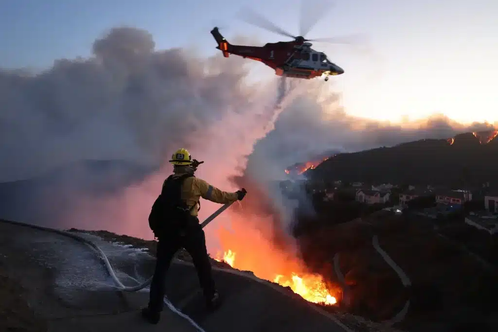 Firefighter spraying water at fire while a helicopter drops water