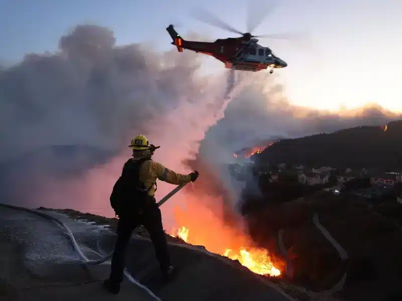 Firefighter spraying water at fire while a helicopter drops water
