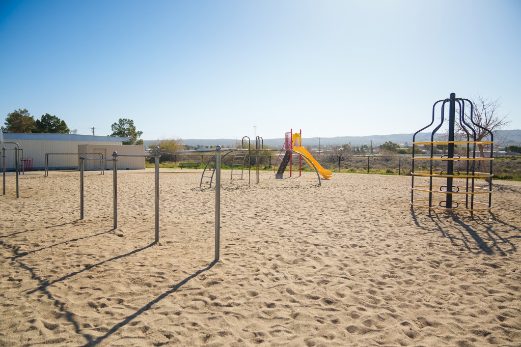 A wide view of playground equipment in the sandbox area of a school playground near a storage unit area. In the background, a wide horizontal view of a nearby city and a mountain range.