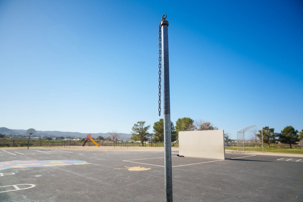 A close-up view of a chain hanging off a pole as part of some playground equipment in the black top area of a school playground. In the background, a wide horizontal view of the sandbox area with more playground equipment, a basketball hoop, and a view of a nearby city and a mountain range.