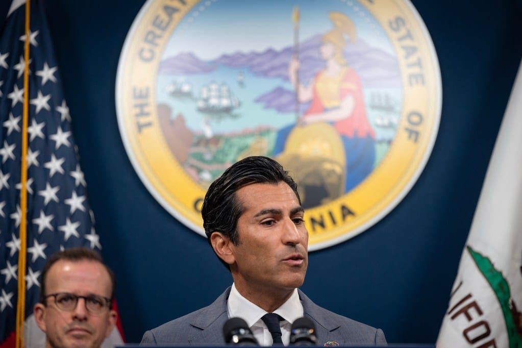 A suited speaker stands at a podium in front of the California state seal, with U.S. and California flags on either side, speaking into microphones while another person listens behind them.